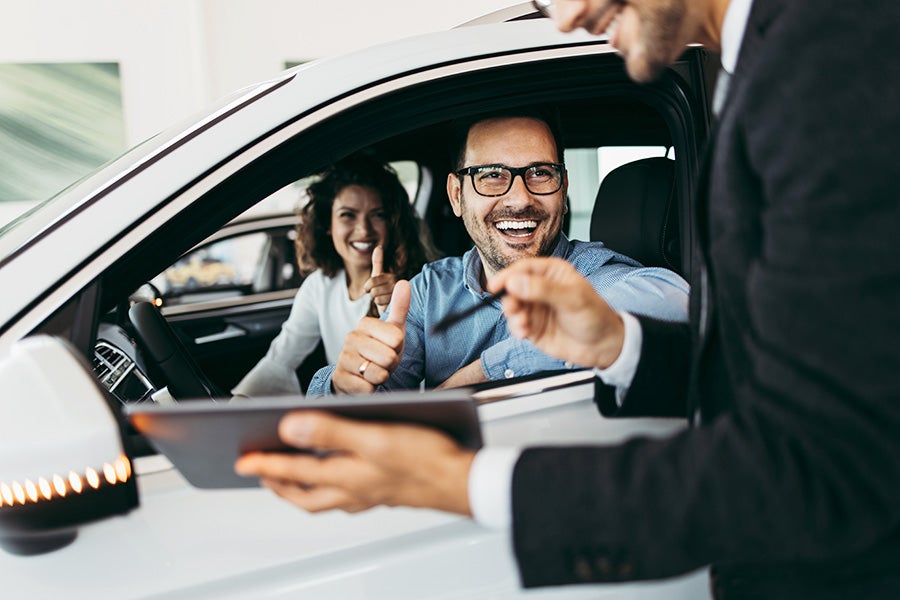 New car financing options for buying a new GMC vehicle is being explained by a sales rep pointing to his IPad to a smiling couple who are sitting in the front seat of the one they like on the sales floor. 